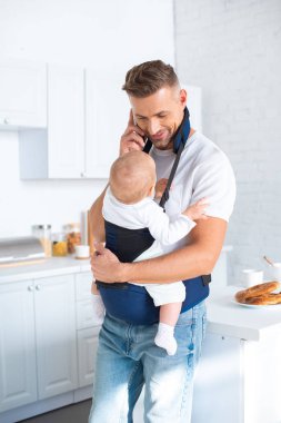 neşeli baba holding bebek kız bebek taşıyıcı ve Smartphone'da konuşuyor 