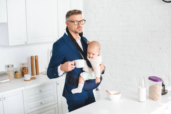handsome businessman holding infant daughter in baby carrier and cup with drink 