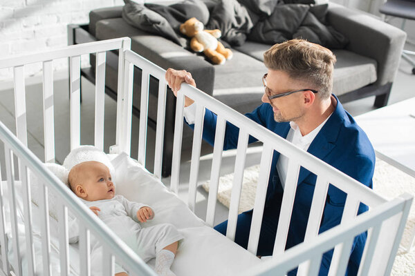 happy father in formal wear sitting near infant daughter in baby crib 