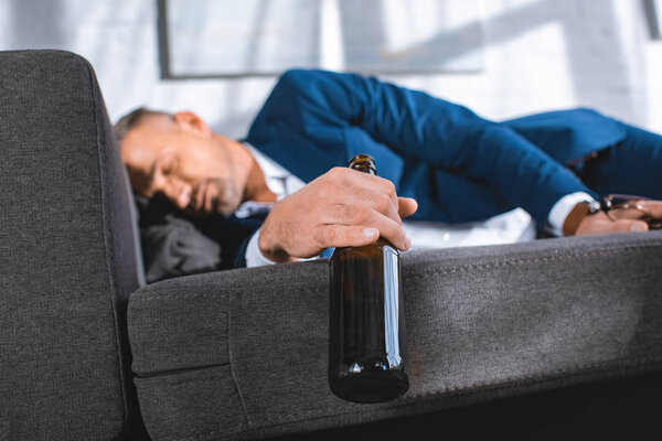 selective focus of bottle in hand of drunk businessman sleeping on sofa