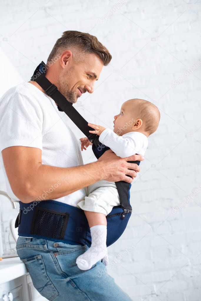 happy father holding infant daughter in baby carrier at home 