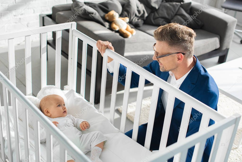 happy father in formal wear sitting near infant daughter in baby crib 