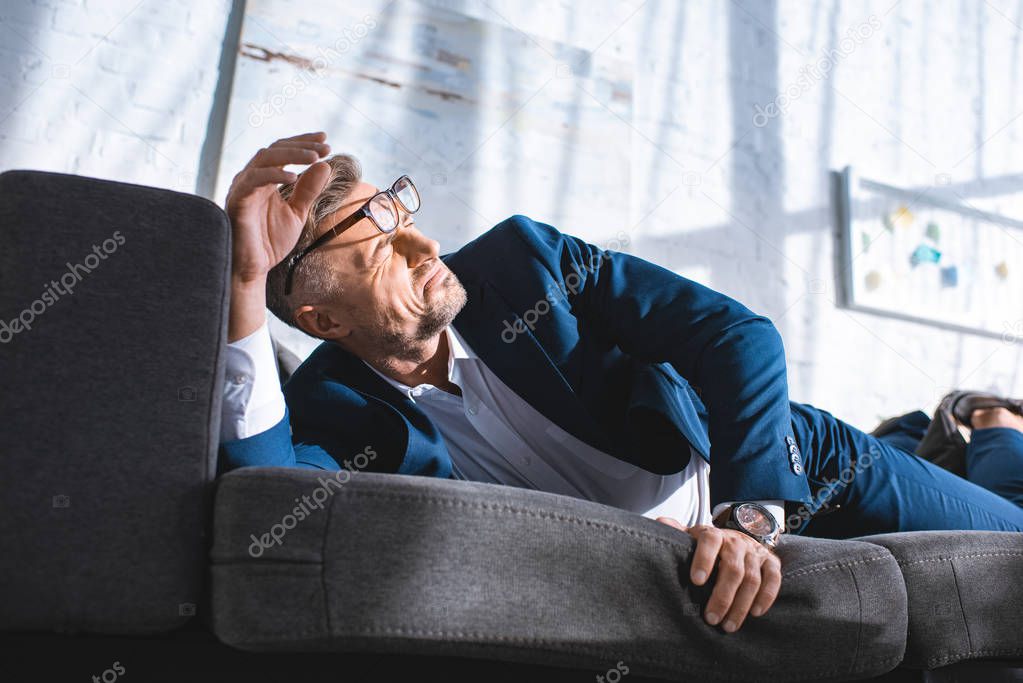 drunk businessman in glasses lying on sofa with sunshine in living room 