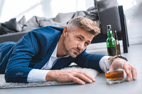избирательный фокус пьяного бизнесмена лежащего на ковре рядом со стеклом с алкогольным напитком
