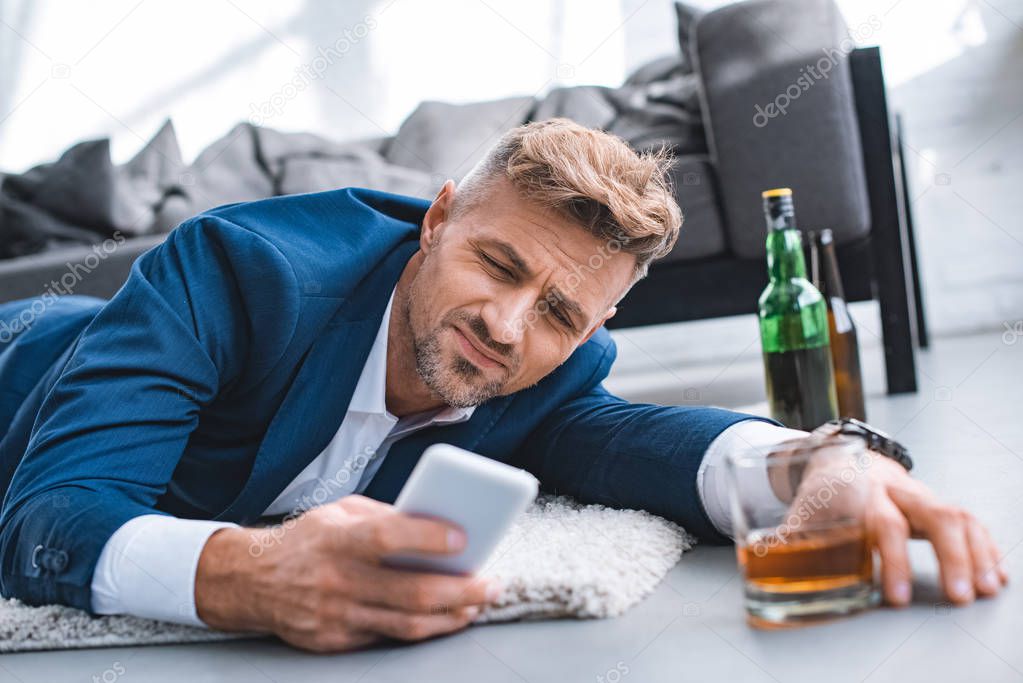 selective focus of drunk businessman lying on carpet and using smartphone  