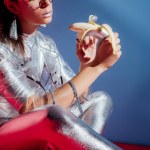 Cropped view of fashionable young woman in metallic bodysuit and raincoat posing with banana on blue background