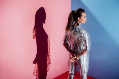 rear view of stylish girl posing in silver bodysuit and trendy raincoat on pink and blue background clipart
