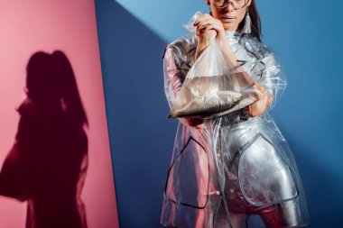 gümüş elbise ve balık pembe ve mavi arka plan üzerinde plastik torba tutarak yağmurluk içinde şık genç kadın kırpılmış görünümü