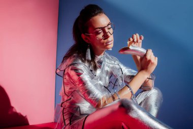 fashionable model in metallic bodysuit and raincoat posing with silver banana on pink and blue background clipart