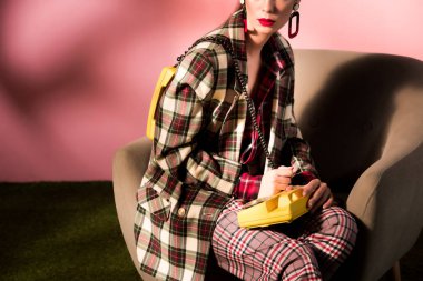 cropped view of girl in checkered suit posing in armchair with vintage telephone on pink background clipart