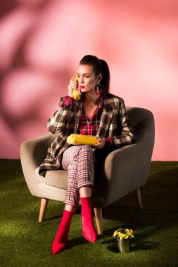 fashionable girl in checkered suit posing in armchair with rotary phone on pink background clipart