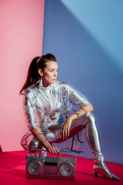 fashionable young woman in silver bodysuit and raincoat posing with boombox on pink and blue background