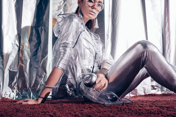 stylish young woman in silver bodysuit and raincoat holding silver apple and posing for fashion shoot on metallic background