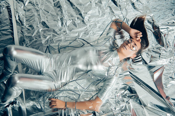 top view of stylish woman posing in silver bodysuit and raincoat on metallic background