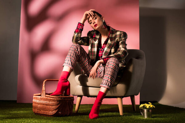 красивая элегантная девушка в клетчатом костюме позирует в кресле с корзиной на розовом фоне
