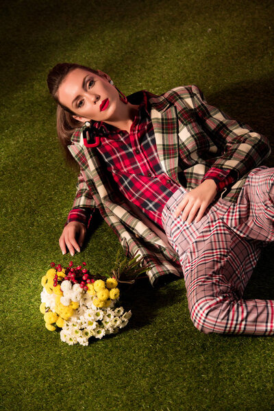 элегантная стильная девушка в осеннем клетчатом костюме позирует с цветами на зеленом ковре
