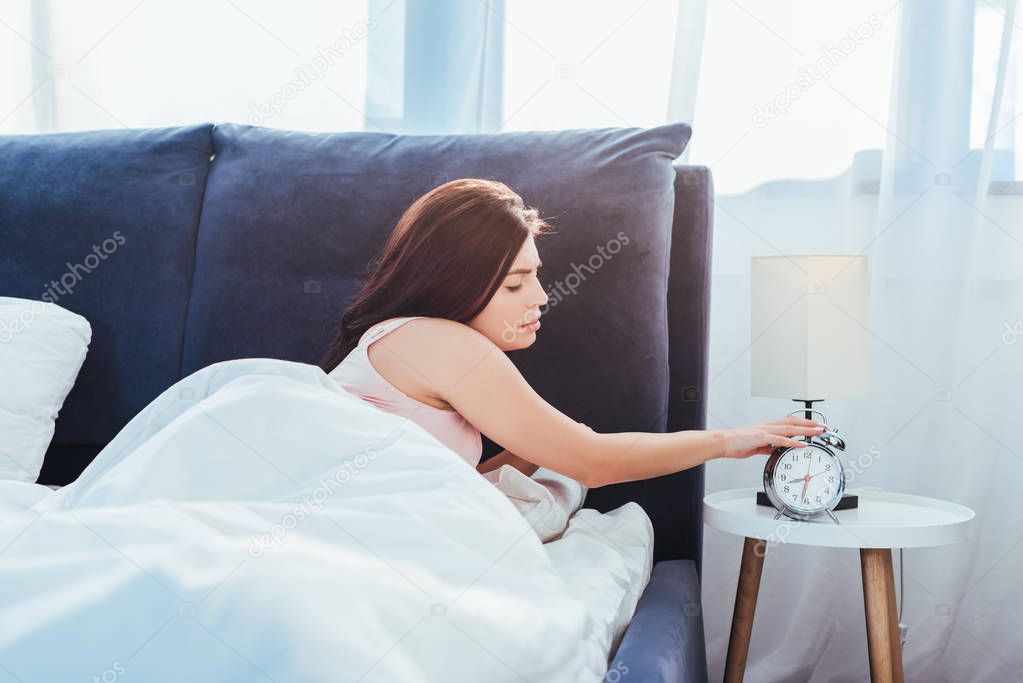 side view of young woman turning off alarm clock in bedroom during morning time at home