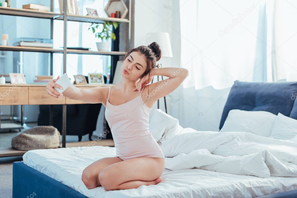 attractive girl taking selfie on smartphone while sitting on bed during morning time at home