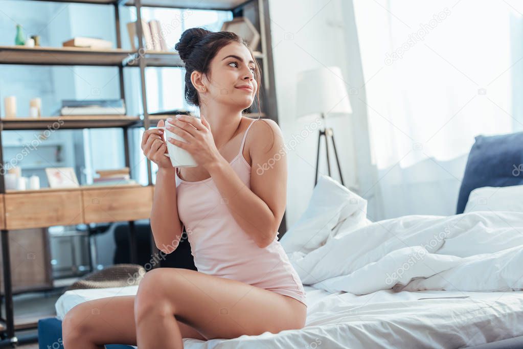 happy young woman sitting with coffee cup on bed during morning time at home