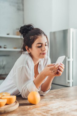 tahta masada portakal evde mutfak ile akıllı telefon kullanan mutlu genç kadın