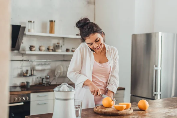 laughing young woman cutting orange by knife and talking on smartphone in kitchen at home