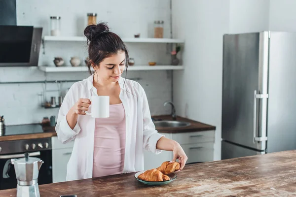 自宅の台所で木製のテーブルでクロワッサンとコーヒーの朝食を持っている陽気な若い女性  — 無料ストックフォト