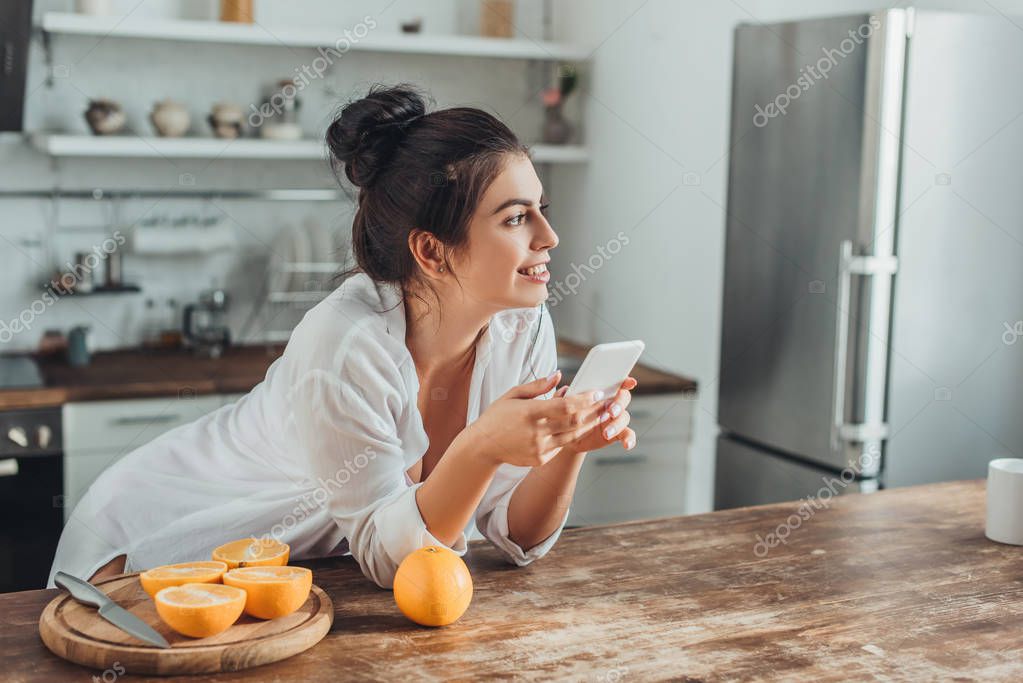 Cheerful young woman using smartphone at wooden table with oranges in kitchen at home