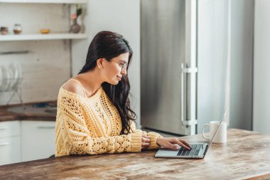 gözlük ile evde mutfak laptop ve kahve Kupası tahta masada oturan kadın freelancer yan görünüm