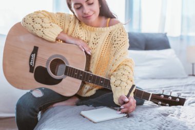 selective focus of girl sitting with acoustic guitar and writing song in textbook on bed at home