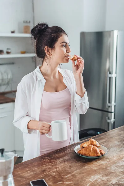自宅の台所で木製のテーブルでクロワッサンとコーヒーの朝食を持つ魅力的な女性  — 無料ストックフォト