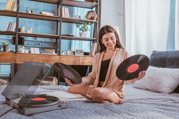 smiling girl holding vinyl record near phonograph and sitting on bed at home