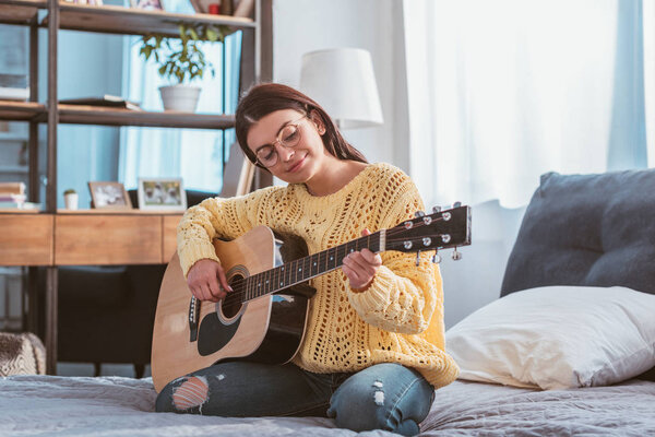 Улыбающаяся красивая женщина в очках, играющая на акустической гитаре, сидя дома на кровати
