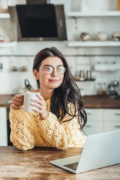 愉快的女性自由职业者在眼镜与咖啡杯坐在木桌与笔记本电脑在厨房在家里 — 图库照片