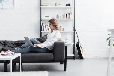 kanepede oturan ve oturma odasında dizüstü kullanarak odaklı kız