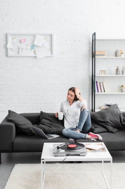 kahve fincanı ile kanepede oturan, dizüstü bilgisayar kullanarak ve vinil plak çalar oturma odasında müzik kız