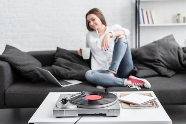 kız beyaz gömlekli kahve fincanı ile kanepede oturan, dizüstü bilgisayar kullanarak ve vinil plak çalar oturma odasında dinleme