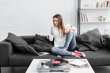 kahve fincanı ile kanepede oturan, dizüstü bilgisayar kullanarak ve vinil plak çalar oturma odasında dinlerken beyaz gömlekli güzel kız