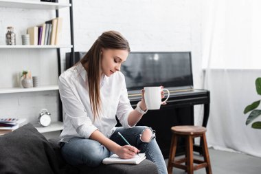 girl sitting with coffee cup on couch and writing in notebook with piano on background clipart