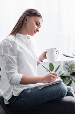 kahve fincanı ile oturan ve evde not defterinde yazılı beyaz gömlekli çekici kız