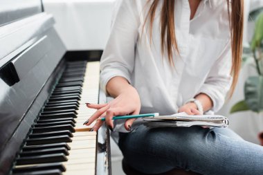 cropped view of girl in white shirt with notebook playing piano and composing music at home clipart