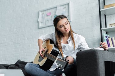gitar ile kanepede oturan, not defterinde yazılı ve oturma odasında müzik bestelemek güzel kız
