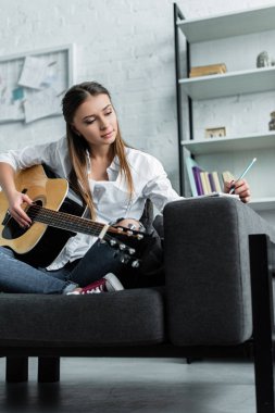 gitar ile kanepede oturan, not defterinde yazılı ve oturma odasında müzik bestelemek dalgın kız