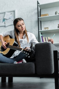 pensive girl sitting on couch with guitar, writing in notebook and composing music in living room clipart