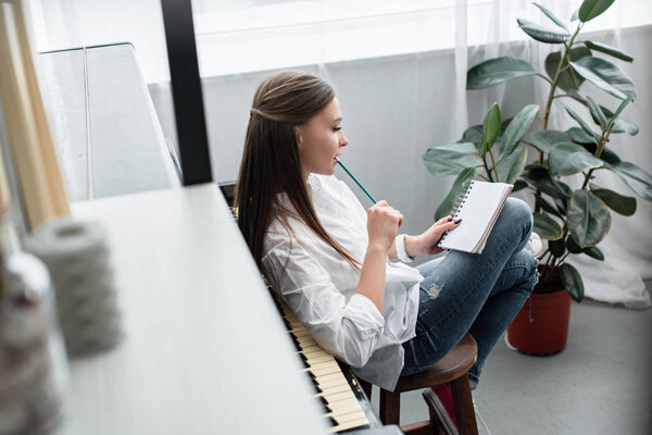 girl with notebook sitting near piano and biting pencil while composing music in living room