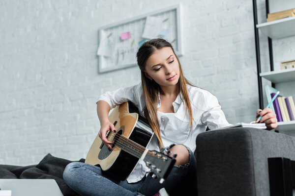 beautiful girl sitting on couch with guitar, writing in notebook and composing music in living room