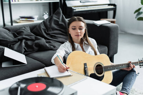 selective focus of girl with acoustic guitar writing in notebook while composing music at home