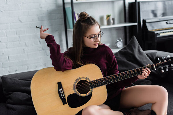 attractive girl sitting, holding marijuana joint and playing guitar at home