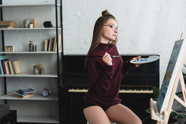 beautiful girl in burgundy sweater sitting, holding palette and painting at home 