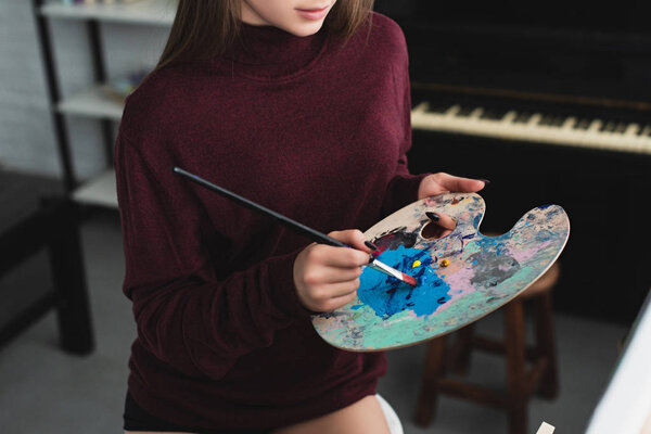partial view of girl in burgundy sweater holding paintbrush with palette and painting at home 