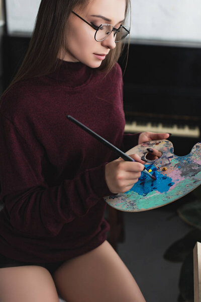 красивая девушка в бордовом свитере сидит, держа кисть, палитру и живопись дома
 